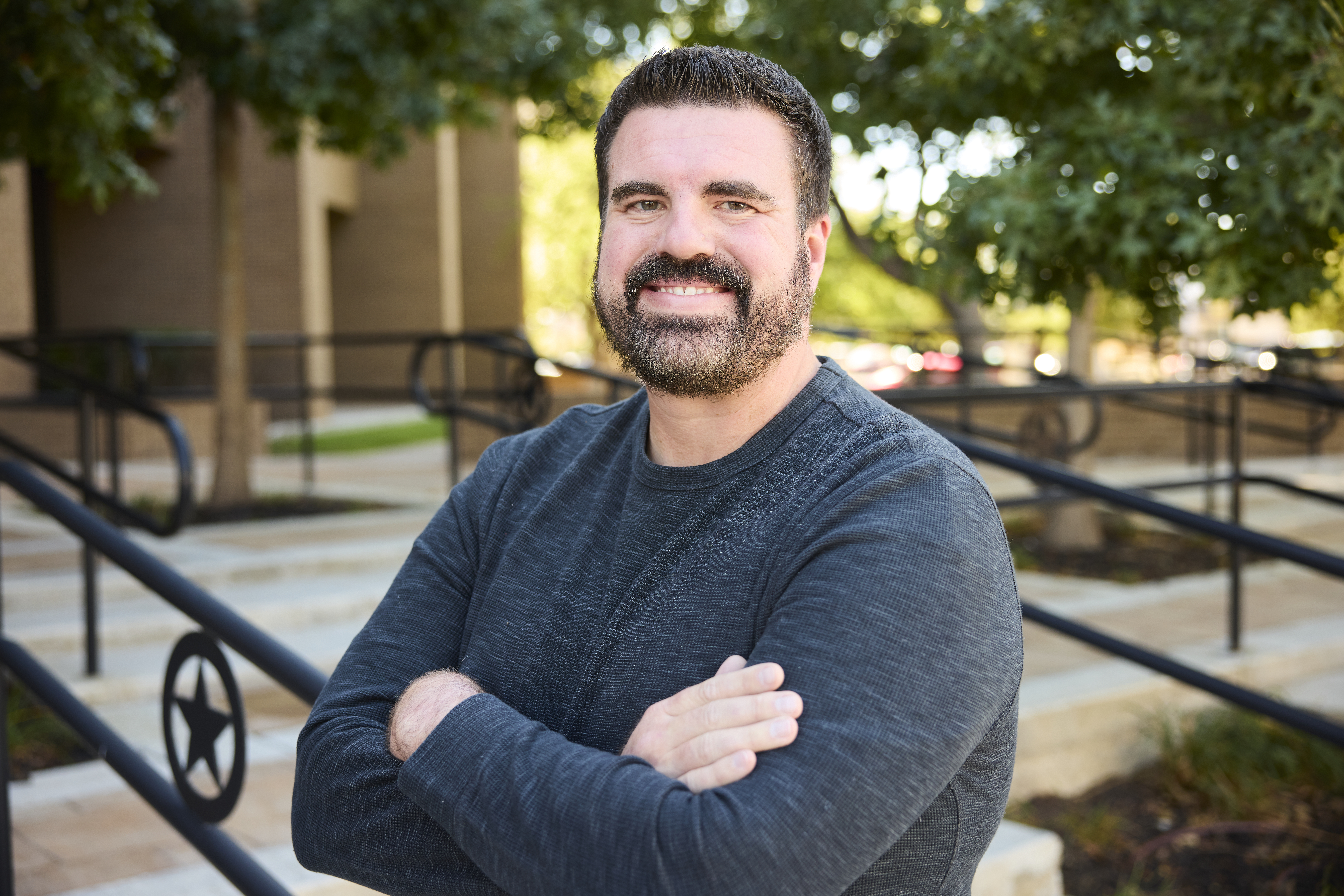 Pantexan Taylor Cockrell A man with dark, short-cut hair and trimmed beard is outside smiling, leaning up against the railing of steps leading up to a building. He’s wearing a dark gray shirt, arms crossed. A few trees with green leaves are in the back ground.