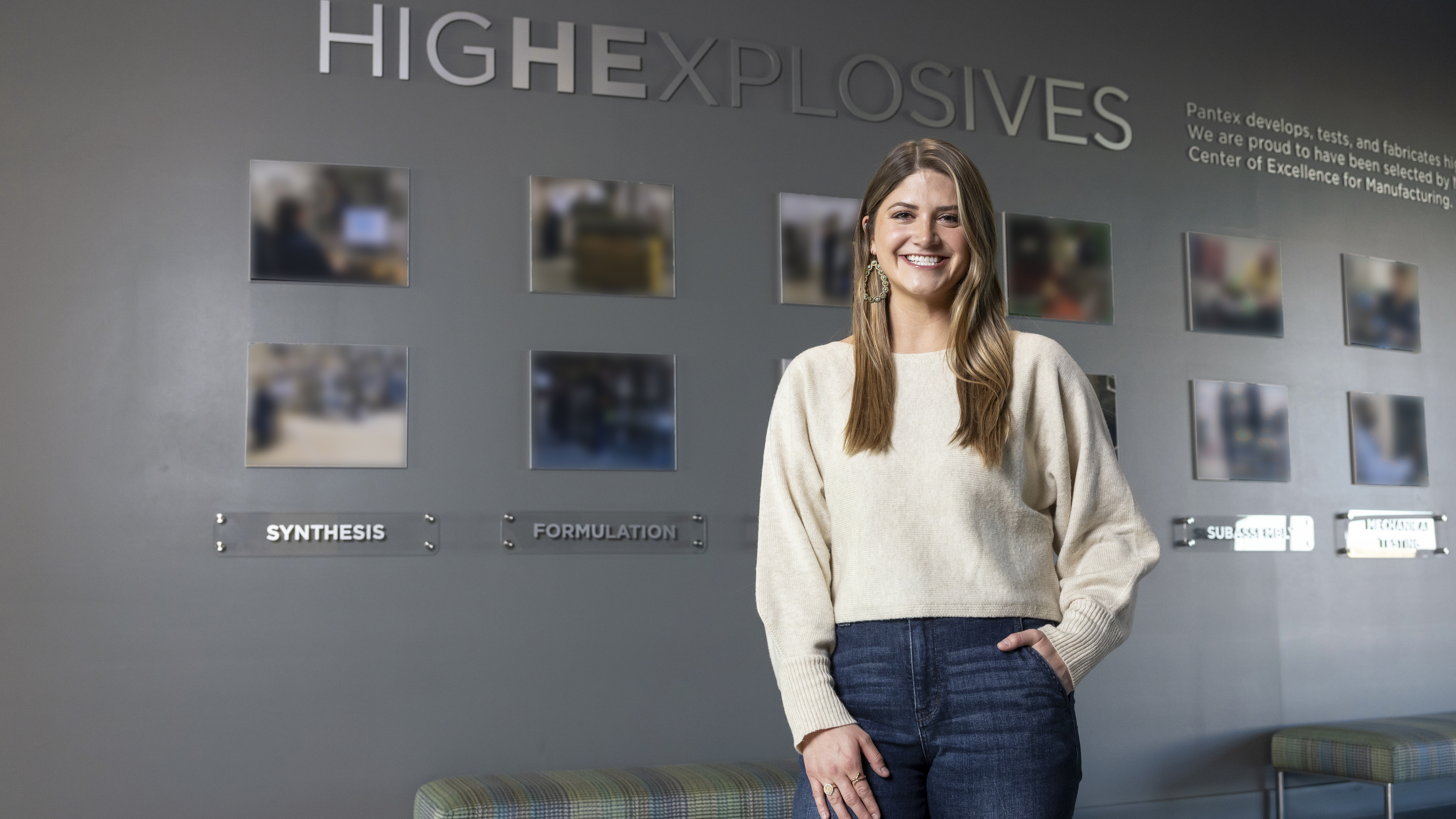 Kennedy Venneman A young blonde woman stands in front of a grey wall lined with photographs. Above her, the words “high explosives” are visible in silver text.