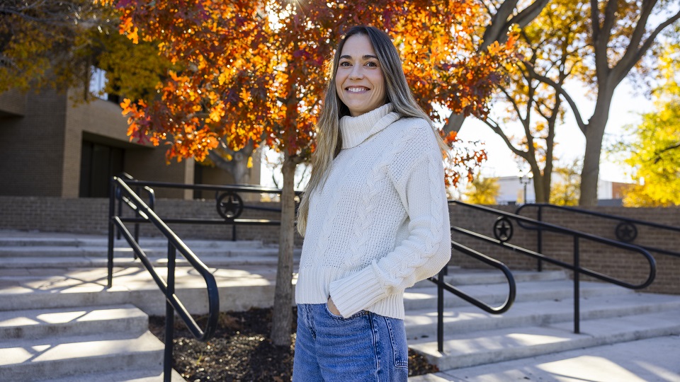 Tiffany Wiley. Photo by Adam Baker A long-haired woman wearing a white turtleneck sweater and blue jeans stand in front of a set of stairs leading into a building. She is turned slightly to her right, hands in pockets. Behind her is a tree full of orange-red leaves.