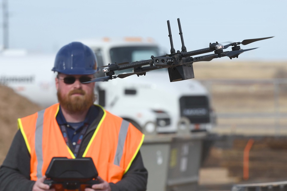 A drone is seen clearly with a man in a construction vest and a hard hat operating the drone is seen blurred behind.