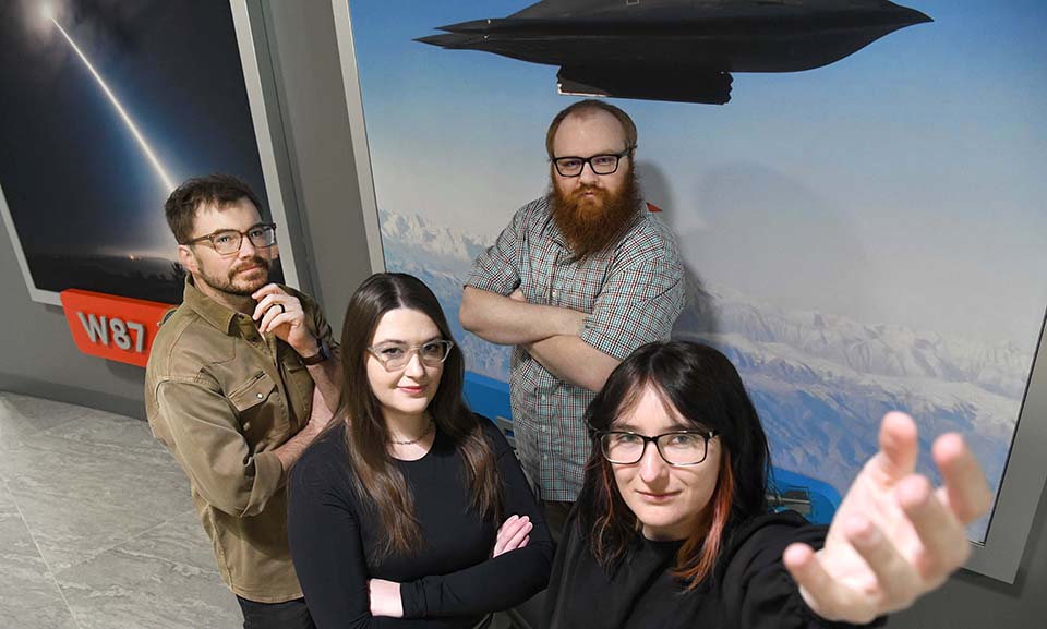  Four individuals stand close together, look up at the camera. First person is close to the camera, wearing a black top and dark-rimmed glasses, hand open, palm up toward the camera.; shoulder length black hair with copper face-framing highlights. Second person is on the left, wearing a black long-sleeved top and glasses, translucent frames. Dark brown hair down to the upper torse, and left arm bent, fingers on arm of glasses as though adjusting them. Third person on left side of photo is wearing a dark tan