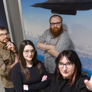 Four individuals stand close together, look up at the camera. First person is close to the camera, wearing a black top and dark-rimmed glasses, hand open, palm up toward the camera.; shoulder length black hair with copper face-framing highlights. Second person is on the left, wearing a black long-sleeved top and glasses, translucent frames. Dark brown hair down to the upper torse, and left arm bent, fingers on arm of glasses as though adjusting them. Third person on left side of photo is wearing a dark tan