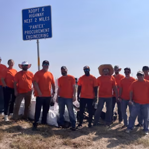 A group of men and women wearing bright orange T-shirts stand in front of a large pile of white trash bags under a road sign reading, “Adopt-A-Highway next two miles. Pantex Procurement Engineering.”
